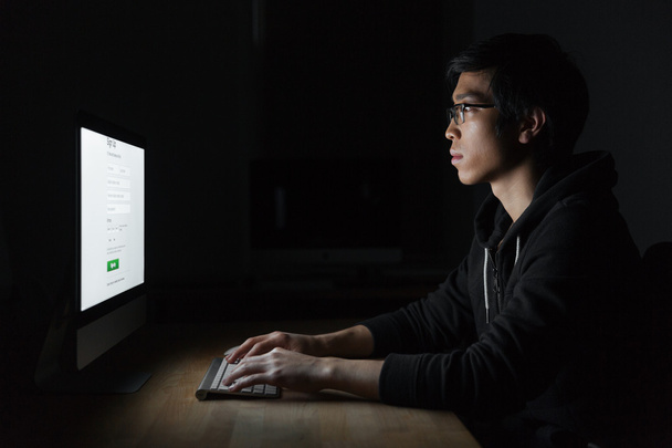 Concentrated asian young man using computer in evening at home - Foto, afbeelding