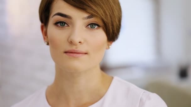 Young beautiful woman with stylish short haircut and fresh make-up looking at camera, indoors - Filmati, video