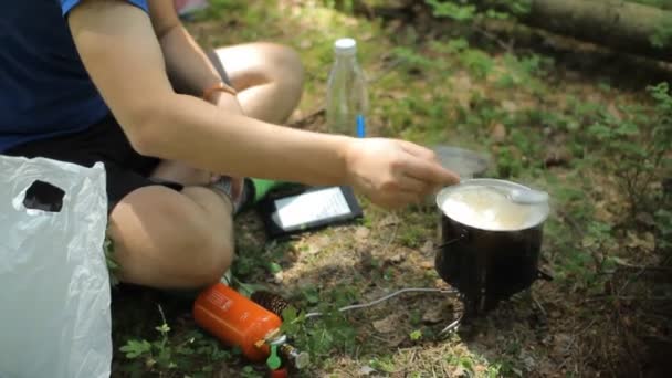 森のガスバーナーの鍋で食事を準備している男。地面に座ってスープをかき混ぜる - 映像、動画