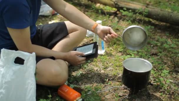 Man reads an e-book in the forest. Near the tourist pot on a gas burner - Filmagem, Vídeo