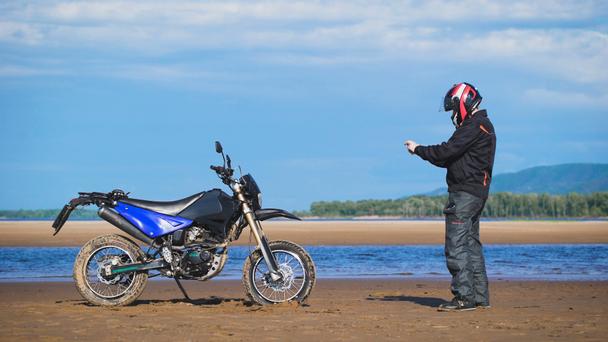 Fietser op de oever van de rivier, naast zijn motorfiets, maakt de foto op de telefoon. - Video