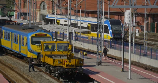 Женщина на платформенном поезде переходит дорогу и садится на локомотив Два синих и желтых пассажирских электрички стоят на железнодорожном вокзале
 - Кадры, видео