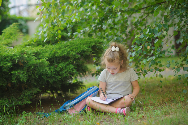 La piccola scolaretta si siede avendo attraversato gambe sotto un albero e fa i compiti
. - Foto, immagini