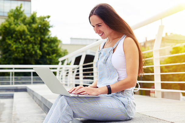  Ciemnowłosa kobieta na nasłoneczniej ulicy z laptopem na kolanach - Zdjęcie, obraz