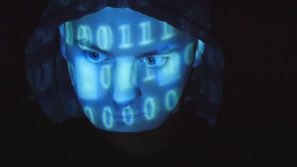 Un hacker incappucciato che lavora su un computer, codice binario proiettato sulla sua faccia. Codice sorgente proiettato su un volto arrabbiato ostile mans, sfondo nero
. - Filmati, video