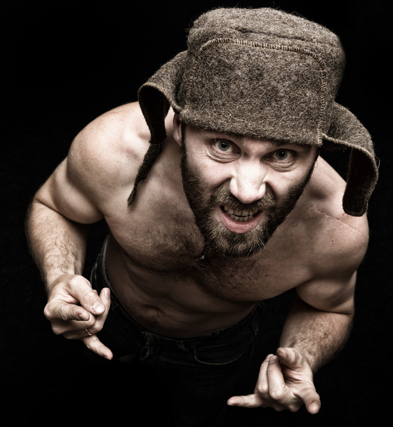 Dark portrait of scary evil sinister bearded man with smirk, makes various hands signs and expresses different emotions. strange Russian man with a naked torso and a woolen hat - Foto, imagen