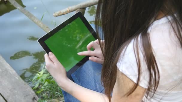 Mãos femininas usando tablet pc digital com tela verde sentado em um molhe de madeira junto ao lago
. - Filmagem, Vídeo