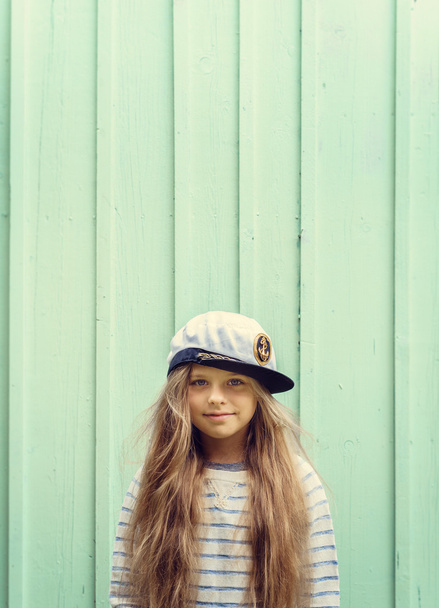 Cute little girl stands near a turquoise wall in sailor hat and smiling Space for text. - Photo, Image