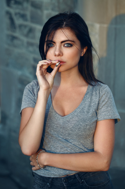 Πορτρέτο του λευκό Καυκάσιος όμορφη σέξι μελαχρινή νεαρή γυναίκα με φωτός μπλε πράσινα μάτια με τζιν και t-shirt καπνίζοντας τσιγάρο ψάχνει κεκλεισμένων των θυρών, τονισμένα με τα φίλτρα του Instagram, απλό τρόπο ζωής - Φωτογραφία, εικόνα