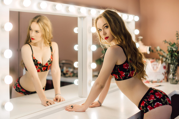 Молода красива жінка наносить макіяж обличчям пензлем, дивлячись у дзеркало, сидить на стільці в роздягальні з вінтажним дзеркалом темної кімнати, в червоній білизні
 - Фото, зображення