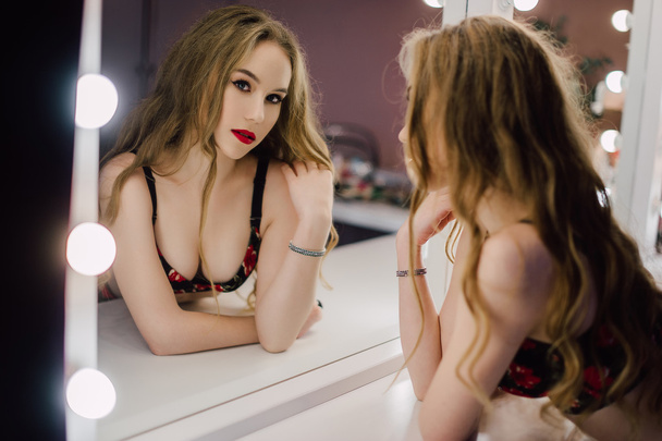Joven hermosa mujer aplicando su maquillaje cara con cepillo, mirando en un espejo, sentado en la silla en el vestidor con espejo vintage habitación oscura, usando lencería roja
 - Foto, imagen