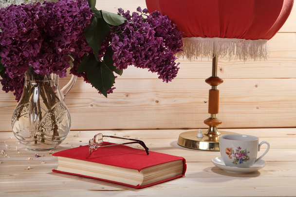 Bouquet of lilacs, book, spectacles, cup of tea and table lamp - Fotó, kép