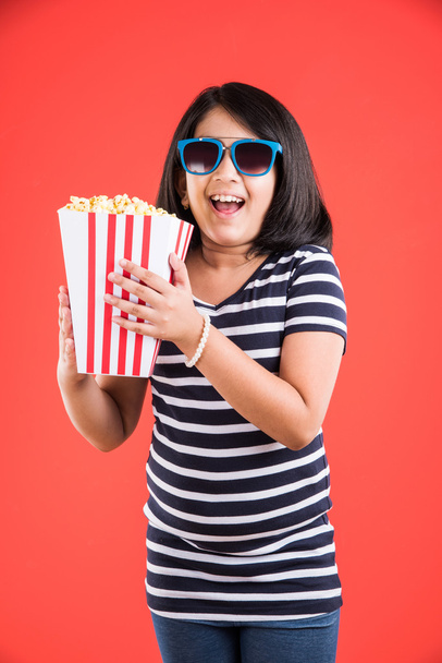 glückliches Mädchen, das Popcorn isst und Gläser trägt, indisches Mädchen, das Popcorn isst, asiatisches Mädchen und Popcorn, kleines Mädchen, das Popcorn auf rotem Hintergrund isst - Foto, Bild