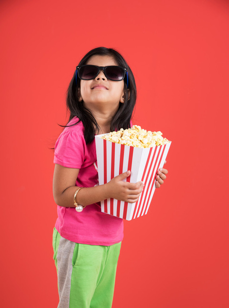 happy girl eating popcorn and wearing glasses, indian girl eating popcorn, asian girl and popcorn, small girl eating popcorn on red background - Foto, afbeelding