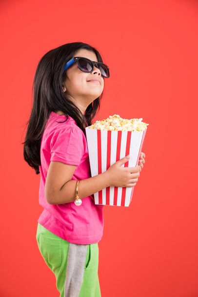 happy girl eating popcorn and wearing glasses, indian girl eating popcorn, asian girl and popcorn, small girl eating popcorn on red background - Valokuva, kuva
