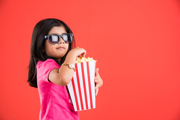 щаслива дівчина їсть попкорн і в окулярах, індійська дівчина їсть попкорн, азіатська дівчина і попкорн, маленька дівчинка їсть попкорн на червоному тлі
 - Фото, зображення