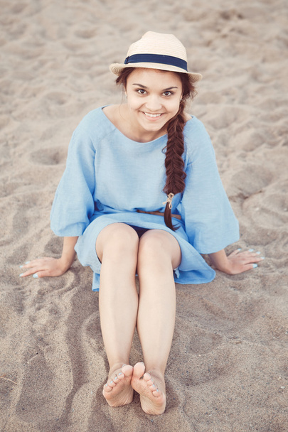 Portré a mosolygó fehér kaukázusi barna nő cserzett bőr kék ruha és a homokos strand partján, naplemente a nyári nap, ülő életmód fogalma szalmakalap - Fotó, kép