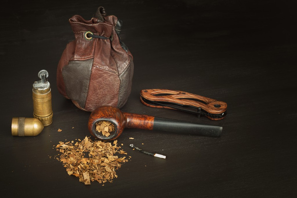 Old tobacco pipe and spilled tobacco, used on a black wooden background. Shabby old tobacco pipe. Wooden tobacco pipe on a black background. Relaxing with a tobacco pipe. Quiet place. Chain smoker. - Fotó, kép