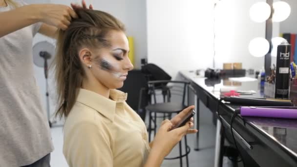 Girl uses smart phone in fashion studio while hairdresser work on clients hair - Séquence, vidéo