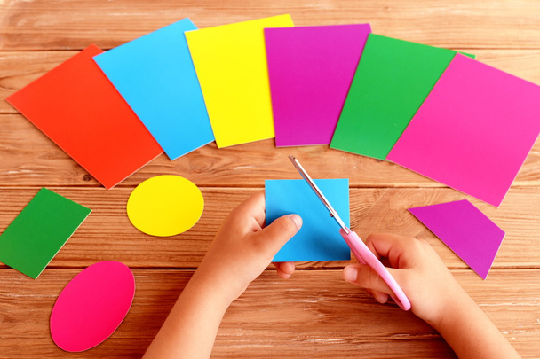 Çocuk makas ve karton kare elinde tutar. Renkli karton yaprak ve ahşap bir masa üzerinde farklı geometrik figürler. Kağıt şekiller kesmek çocuk öğrenir. Erken çocukluk gelişimi  - Fotoğraf, Görsel