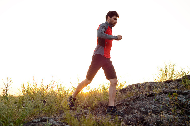 Νεαρός αθλητής τρέχει στο βραχώδες βουνό ίχνος στο ηλιοβασίλεμα. Δραστήριο τρόπο ζωής - Φωτογραφία, εικόνα