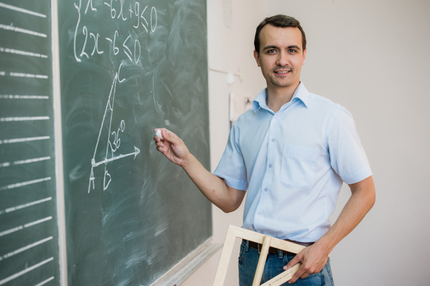 Jonge leraar of student bedrijf driehoek wijzend op schoolbord met formule, op zoek naar camera en glimlachen - Foto, afbeelding