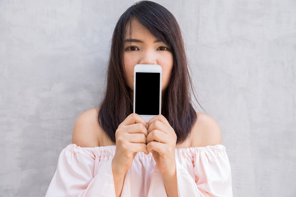Улыбающаяся женщина показывает чистый экран смартфона, стоящий на бетонной стене
. - Фото, изображение