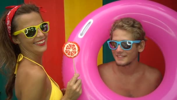 Funny young happy couple dancing isolated on colorful wall in slow motion. Girl is licking orange lollipop and boy is holding pink inflatable ring. - Footage, Video