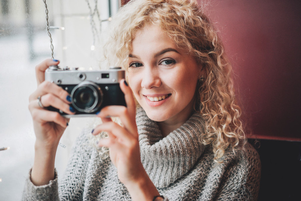 Young blond curly female in warm sweater with old film camera smiling and shooting a photo in the cafe, winter city outside the window - Photo, Image