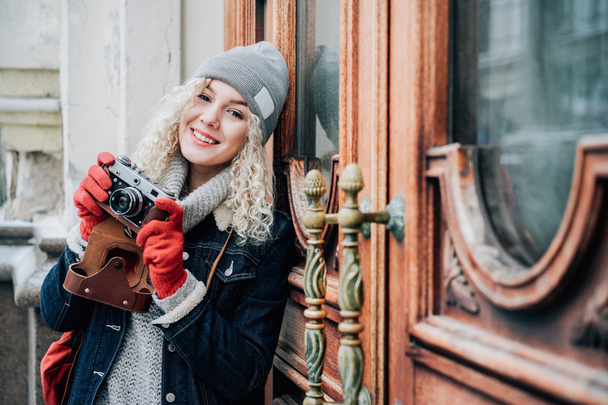 Young blond curly female in warm clothes with old film camera shooting a photo on the background of wooden doors with handles, winter - Photo, Image