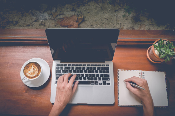 Бизнесмен с ноутбуком и ручкой с блокнотом и чашкой кофе с латте-артом на вид сверху с эффектом винтажного фильтра
 - Фото, изображение