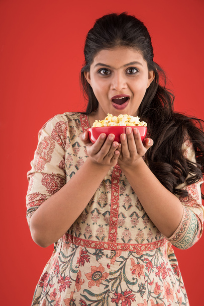 indio joven y hermosa chica comiendo palomitas de maíz, chica asiática disfrutando de palomitas de maíz o maíz pop, aislado sobre fondo rojo
 - Foto, Imagen