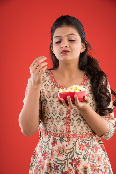 indio joven y hermosa chica comiendo palomitas de maíz, chica asiática disfrutando de palomitas de maíz o maíz pop, aislado sobre fondo rojo
 - Foto, Imagen