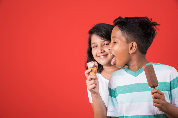 two happy indian kids and ice cream, two asian kids enjoying ice cream or cone or chocolate candy, girl and boy eating ice cream, isolated on red background, - Fotó, kép