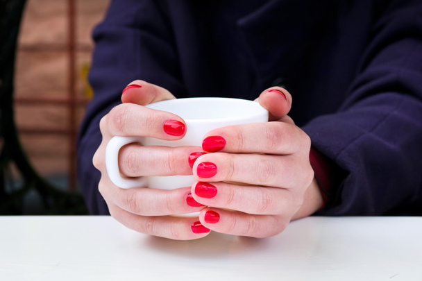 Mujer con esmalte de uñas rojo sosteniendo taza blanca en un café al aire libre
 - Foto, imagen