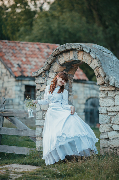 Beautiful girl in white vintage dress walking near ancient stone house. Vintage bridal concept - Foto, imagen