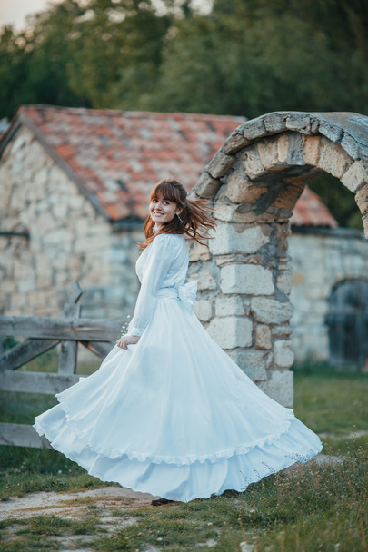 Beautiful girl in white vintage dress walking near ancient stone house. Vintage bridal concept - Фото, изображение