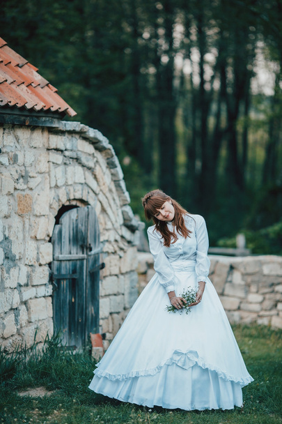 Beautiful girl in white vintage dress walking near ancient stone house. Vintage bridal concept - Φωτογραφία, εικόνα