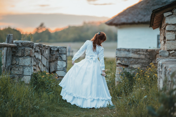 Beautiful girl in white vintage dress walking near ancient stone house. Vintage bridal concept - Fotó, kép
