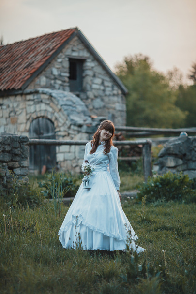 Beautiful girl in white vintage dress walking near ancient stone house. Vintage bridal concept - 写真・画像