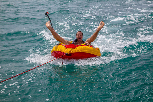 Hombre sentado en anillo inflable remolcado por un barco en el agua y haciendo caras a la cámara y sosteniendo la cámara Go Pro
 - Foto, imagen