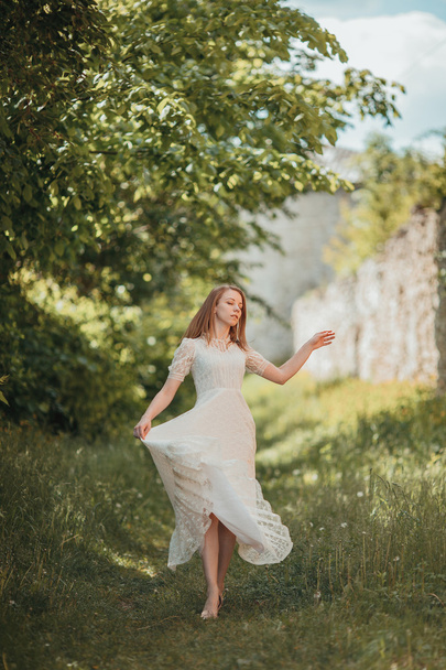 Beautiful young girl in white vintage dress walking near ancient castle. - Фото, изображение