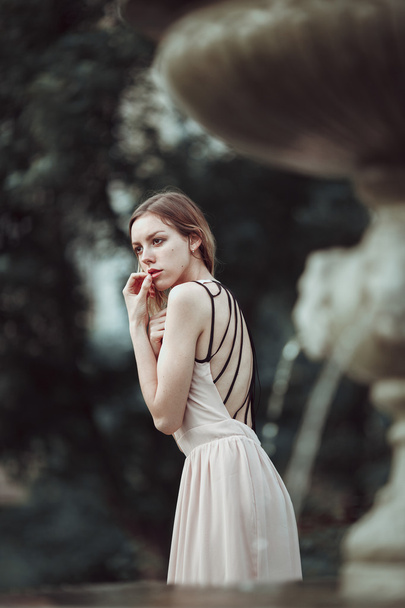 Szép, fiatal lány a divatos rózsaszín ruha álló vintage kő szökőkút közelében. - Fotó, kép