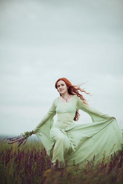 Young redhead girl in medieval dress walking through field with sage flowers. Wind concept. Fantasy - Φωτογραφία, εικόνα
