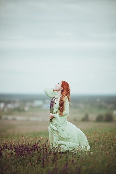 Menina ruiva jovem em vestido medieval andando através do campo com flores de sálvia. Conceito de vento. Fantasia
 - Foto, Imagem