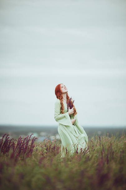 Young redhead girl in medieval dress walking through field with sage flowers. Wind concept. Fantasy - Foto, Bild