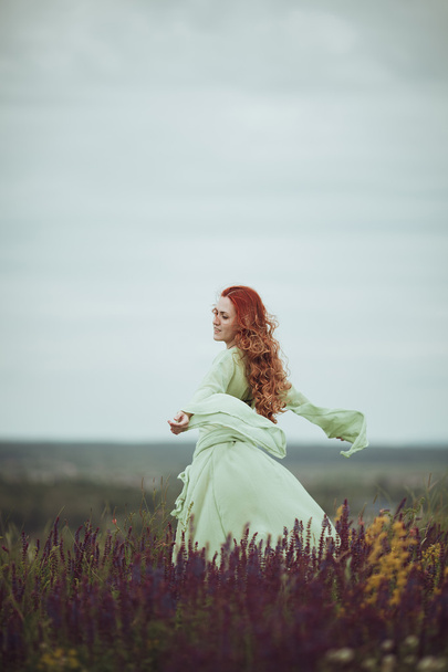 Young redhead girl in medieval dress walking through field with sage flowers. Wind concept. Fantasy - Foto, Bild