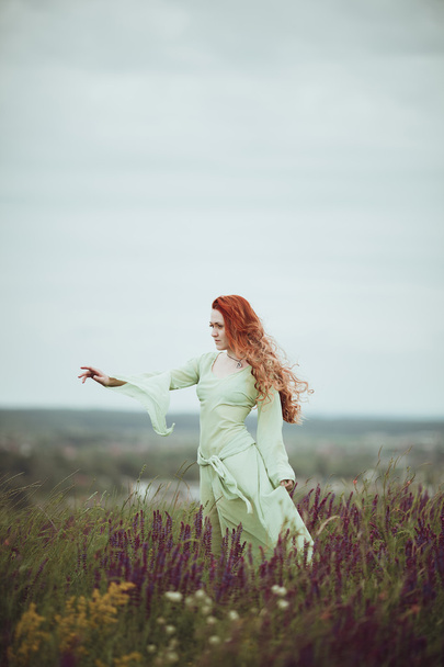 Young redhead girl in medieval dress walking through field with sage flowers. Wind concept. Fantasy - Photo, Image