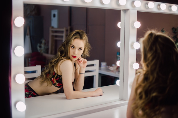 Jeune belle femme appliquant son visage de maquillage avec une brosse, regardant dans un miroir, assise sur une chaise au dressing avec miroir vintage chambre sombre, portant de la lingerie rouge
 - Photo, image