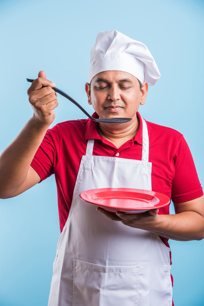 Retrato de um cozinheiro indiano macho chef segurando uma placa vermelha e concha isolada em fundo azul, asiático macho chef degustação
 - Foto, Imagem
